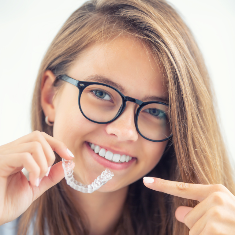 Smiling young woman wearing glasses holding a clear Invisalign aligner and pointing at her teeth, showcasing straight teeth and orthodontic treatment option.